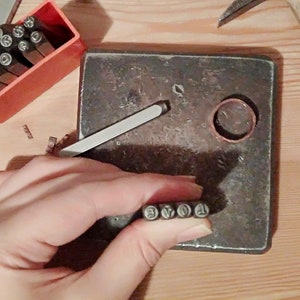 May include: A person's hand holding a set of metal number stamps. The stamps are arranged in order from 1 to 6. The hand is holding the stamps over a metal surface. There is a copper ring and a metal tool in the background.