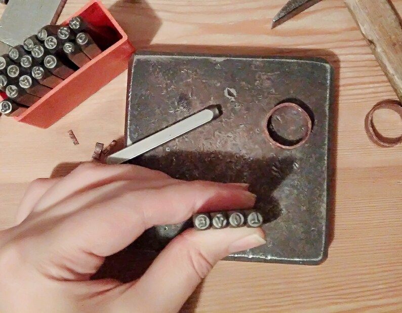 May include: A person's hand holding a set of metal number stamps. The stamps are arranged in order from 1 to 6. The hand is holding the stamps over a metal surface. There is a copper ring and a metal tool in the background.