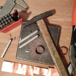 May include: A close-up of a metalworking setup with tools including a hammer, a metal anvil, a pair of metal shears, a metal ruler, and a set of metal stamps in a red plastic case. There is also a piece of copper sheet metal and a metal ring.