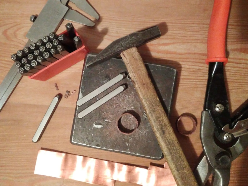 May include: A close-up of a metalworking setup with tools including a hammer, a metal anvil, a pair of metal shears, a metal ruler, and a set of metal stamps in a red plastic case. There is also a piece of copper sheet metal and a metal ring.
