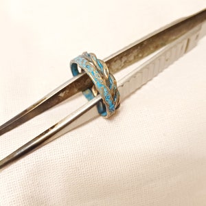 May include: A silver ring with a blue enamel design. The ring is held by a pair of tweezers.
