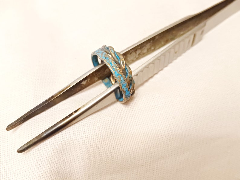 May include: A silver ring with a blue enamel design. The ring is held by a pair of tweezers.