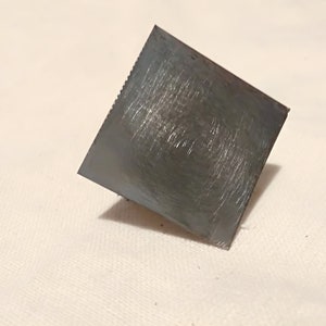 May include: A square, silver-colored metal scraper with a textured surface.