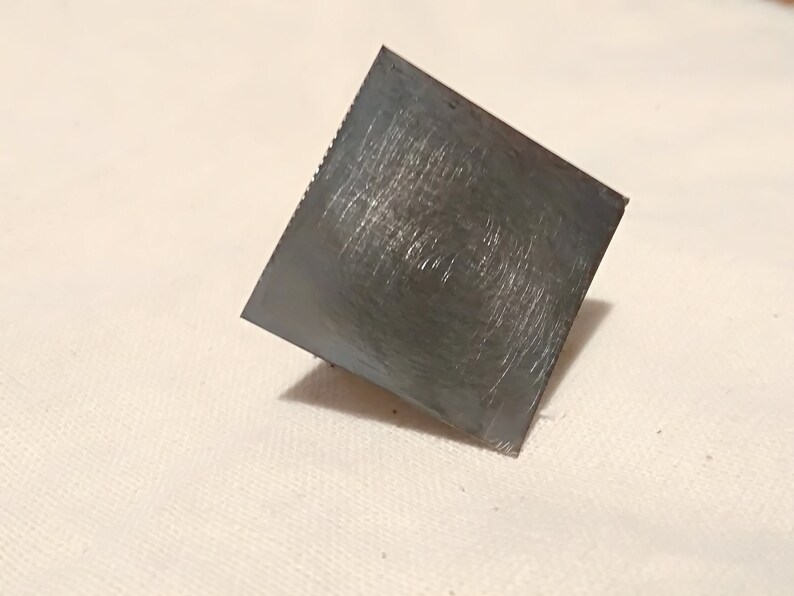 May include: A square, silver-colored metal scraper with a textured surface.