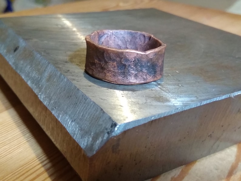 May include: A wide copper ring with a hammered texture. The ring is sitting on a metal surface.