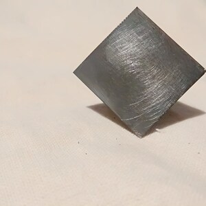 May include: A square piece of metal with a brushed finish. The metal is a light gray color.
