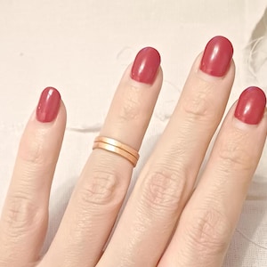 Puede incluir: Una mano con dos finas bandas doradas en el dedo anular. Las uñas están pintadas de un rojo intenso.