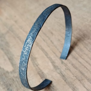 May include: A textured, dark gray metal cuff bracelet. The open-ended bracelet has a slightly hammered appearance. The bracelet is shown on a light brown wooden surface.