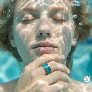 Puede incluir: Un anillo de banda ancha con una incrustación de piedra turquesa vibrante con vetas oscuras naturales. El anillo se lleva en un dedo, sobre un fondo de agua azul brillante con refracciones de luz y pequeñas burbujas de aire visibles.