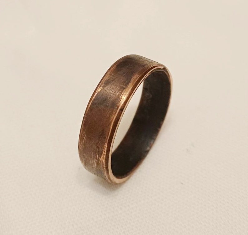 May include: A wide copper band ring with a smooth, slightly textured finish. The ring has a beveled edge and a slightly darker inner band.