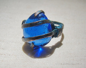 May include: A copper wire ring with a large, blue glass stone set in the centre. The stone is surrounded by copper wire that is wrapped around it.