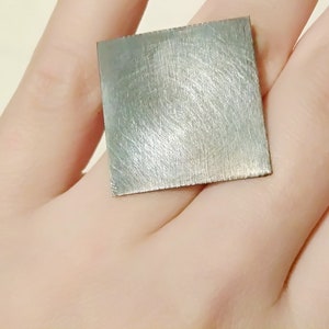 May include: A square piece of brushed metal, possibly silver or aluminum, is held in a hand. The metal has a smooth, brushed finish and appears to be a small, flat piece of metal.