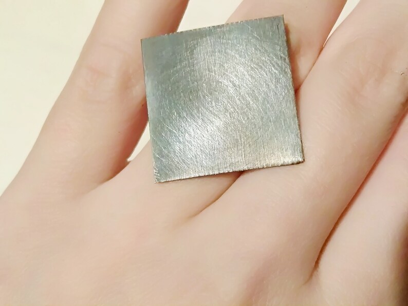 May include: A square piece of brushed metal, possibly silver or aluminum, is held in a hand. The metal has a smooth, brushed finish and appears to be a small, flat piece of metal.