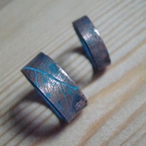 May include: Two copper rings with a blue patina and a leaf design. The rings are on a wooden surface.