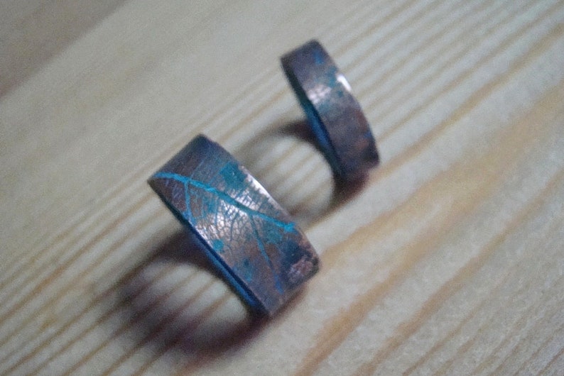 May include: Two copper rings with a blue patina and a leaf design. The rings are on a wooden surface.