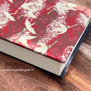 Puede incluir: Un libro rojo con un patrón de grúa blanca en la portada. El libro tiene una columna vertebral blanca con detalles dorados y un borde azul.