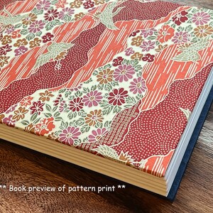 Op de afbeelding: Een boek met een rood, wit en gouden bloemenpatroon op de omslag. Het boek is gesloten en de rug is zichtbaar. Het boek ligt op een houten oppervlak.