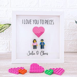 May include: A framed artwork with the text "I LOVE YOU TO PIECES" and the names "Julie & Chris". The artwork features a pink heart and two Lego figures. Several Lego hearts in pink, orange, and green are at the bottom.