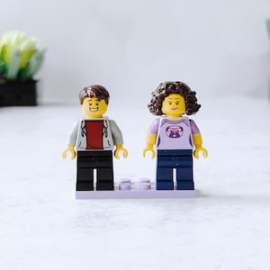 May include: Two Lego minifigures, one with brown hair and a white jacket, the other with brown hair and a purple shirt, standing on a purple base.