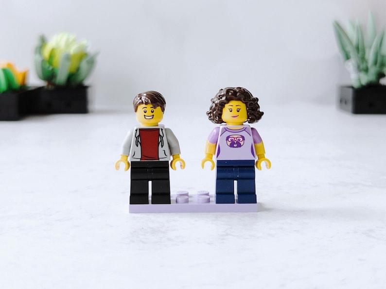 Two mini figures on a lavender base plate. Male has short brown hair, gray and brown shirt and black pants. Female has medium length, curly, brown hair, a purple shirt with a dog wearing red glasses and dark blue pants.