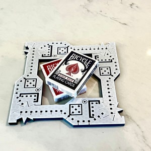 以下が含まれることがあります： 黒色の印と星形のデザインが施された白いプラスチック製のゲームボード。ボードの上にバイシクルのトランプが2組置かれている。一番上のカードはスペードのエースである。