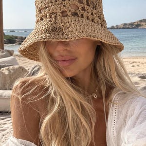 Pode incluir: Um chapéu de crochê bege estilo bucket hat com um padrão de trama aberta e aba larga. O chapéu é feito de fibras em tons naturais, apresentando uma copa estruturada e uma aba flexível inclinada para baixo com uma textura visível.
