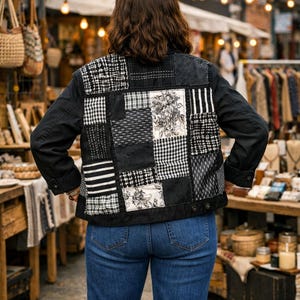 May include: A black jacket featuring a patchwork back panel composed of various black and white fabric squares, including floral, striped, and checkered patterns. The jacket has long sleeves and is worn with blue denim jeans.