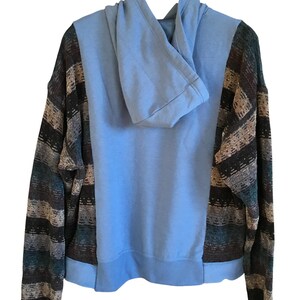 May include: A long-sleeve hoodie featuring a solid light blue center panel and hood, contrasted by sleeves and side panels with a multi-colored horizontal striped pattern in shades of dark brown, tan, black, and muted teal blue.
