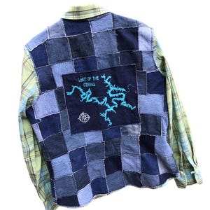 May include: A green and blue plaid long-sleeved shirt with a patchwork denim back. The back features a blue patch with the text "Lake of the Ozarks" and a turquoise outline of a lake.