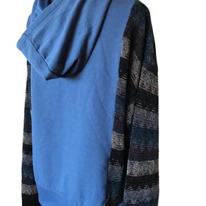 May include: A hooded sweatshirt featuring a solid light blue center panel and hood, contrasted by dark sleeves and side panels with a horizontal knit pattern in shades of black, grey, and teal. The garment has long sleeves and a ribbed cuff.