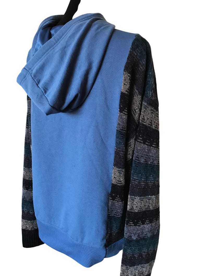 May include: A hooded sweatshirt featuring a solid light blue center panel and hood, contrasted by dark sleeves and side panels with a horizontal knit pattern in shades of black, grey, and teal. The garment has long sleeves and a ribbed cuff.