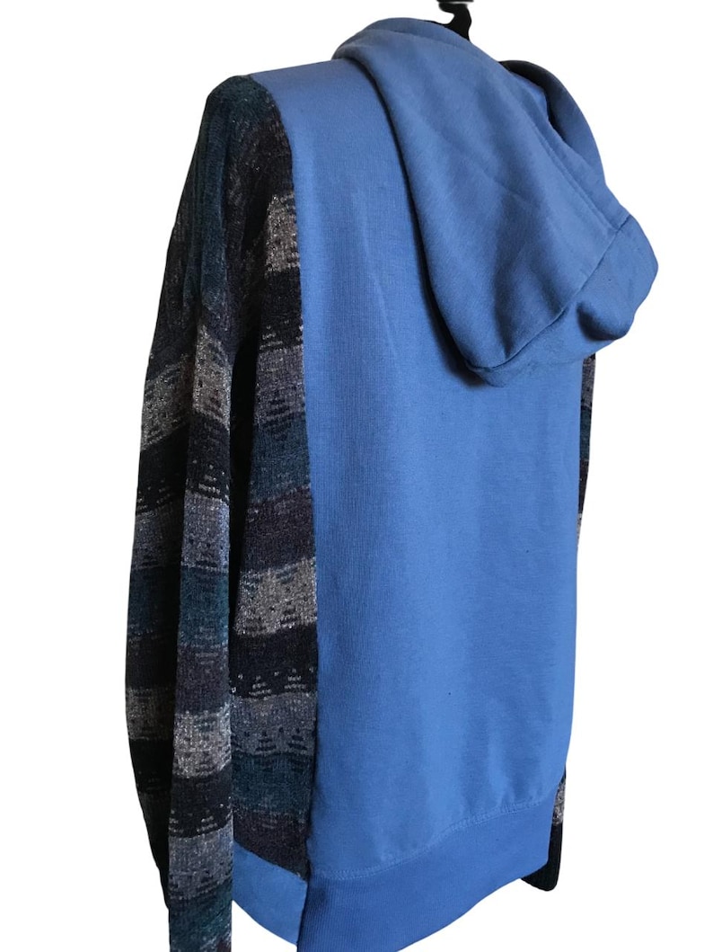 May include: A hooded sweatshirt featuring a solid light blue back and hood, contrasted with long sleeves and side panels displaying a horizontal striped pattern in shades of dark blue, charcoal gray, and light gray with a textured knit appearance.