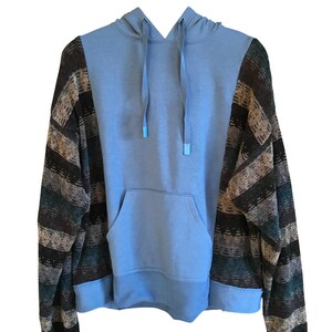 May include: A light blue hoodie featuring long sleeves with a multi-colored horizontal striped pattern in shades of brown, black, and teal. The garment includes a front kangaroo pocket, a hood with matching light blue drawstrings, and ribbed cuffs.