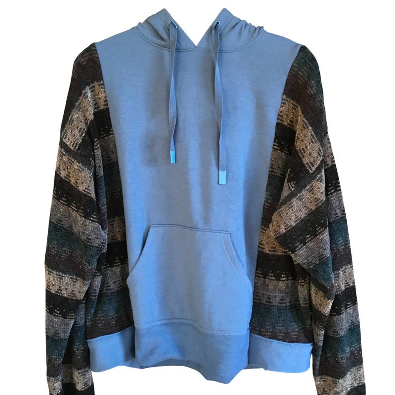 May include: A light blue hoodie featuring long sleeves with a multi-colored horizontal striped pattern in shades of brown, black, and teal. The garment includes a front kangaroo pocket, a hood with matching light blue drawstrings, and ribbed cuffs.