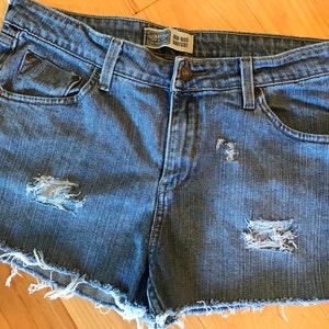 May include: Light blue denim shorts with distressed details. Features include a button closure, front and back pockets, and a raw hem. The shorts have a mid-rise fit and a faded wash.
