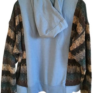 May include: A long-sleeve hooded sweatshirt featuring a solid light blue center panel and hood, contrasted by side panels and sleeves with a horizontal striped pattern in shades of dark brown, tan, teal, and black with a ribbed hem and cuffs.