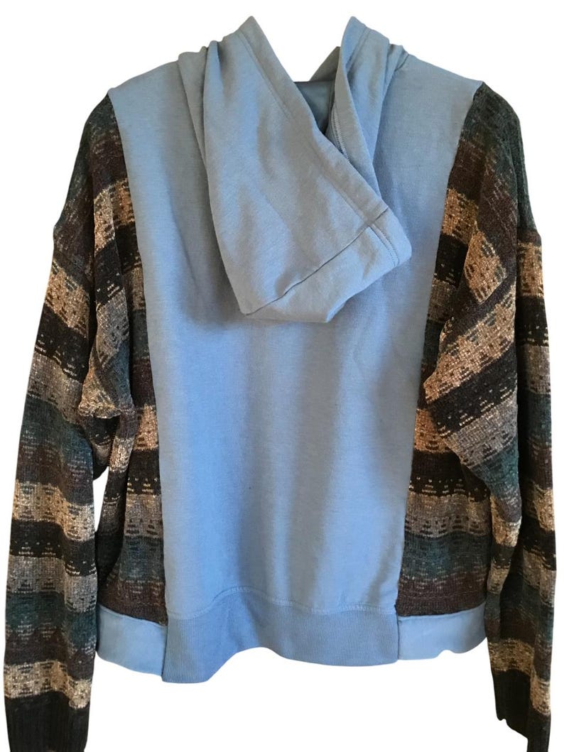 May include: A long-sleeve hooded sweatshirt featuring a solid light blue center panel and hood, contrasted by side panels and sleeves with a horizontal striped pattern in shades of dark brown, tan, teal, and black with a ribbed hem and cuffs.