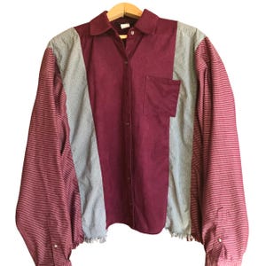 May include: A long-sleeved button-up shirt with a patchwork design. The shirt is made of three different fabrics: a burgundy corduroy, a light gray fabric with a subtle texture, and a red and white striped fabric. The shirt has a frayed hem and a single pocket on the left side.