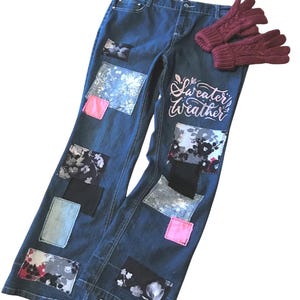May include: Blue denim jeans with decorative patches in various patterns and colors, including floral and solid hues. The jeans feature the text "Sweater Weather" in a cursive font. A pair of burgundy knit gloves rests on top of the jeans.