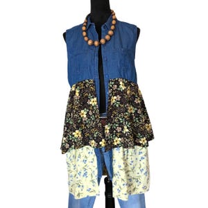 May include: A sleeveless denim vest with a floral patchwork design. The vest features a dark blue denim top, a black floral midsection, and a light yellow floral bottom. A wooden bead necklace is also included.