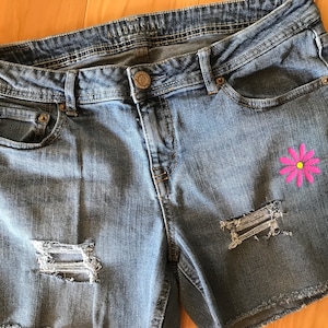 May include: Light blue denim shorts with distressed details and a pink daisy embroidery. The shorts have a classic five-pocket design, a button closure, and the brand name "AEROPOSTALE" on the waistband.