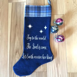 May include: A navy blue Christmas stocking with white text that reads "Joy to the world, The Lord is come. Let Earth receive her king!" The stocking has a plaid cuff and star graphics. Three Christmas ornaments are next to the stocking.
