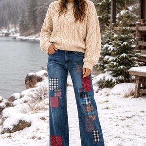 May include: A beige cable-knit sweater, a multi-colored knit beanie, and blue denim jeans featuring various rectangular fabric patches in plaid, checkered, and floral patterns. The jeans have a wide-leg cut and are paired with brown boots.