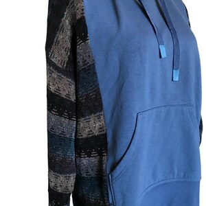 May include: A long-sleeve hoodie featuring a solid blue front and back panel with a kangaroo pocket. The sleeves are constructed from a dark, textured fabric with horizontal stripes in shades of black, grey, and deep blue. It includes a matching blue hood with drawstrings.