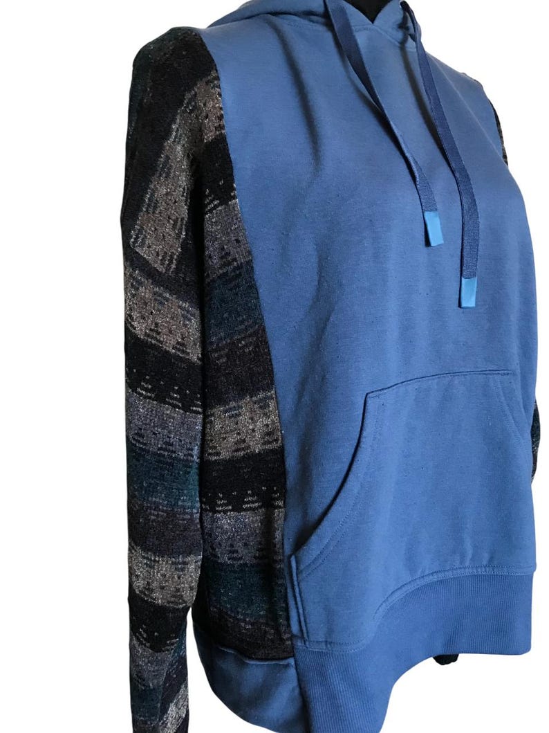 May include: A long-sleeve hoodie featuring a solid blue front and back panel with a kangaroo pocket. The sleeves are constructed from a dark, textured fabric with horizontal stripes in shades of black, grey, and deep blue. It includes a matching blue hood with drawstrings.