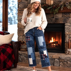 May include: A cream-colored knit sweater, blue denim jeans with various rectangular patches in light blue, dark blue, and red plaid, and white sneakers. The jeans feature a raw hem, and the sweater is paired with a white knit beanie with a pom-pom.