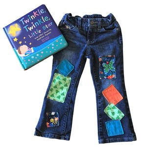 May include: Blue denim jeans with colorful fabric patches sewn on the legs. The jeans have a bootcut style. A book titled "Twinkle, Twinkle Little Star" is in the background.