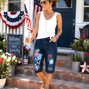 May include: A white sleeveless tank top with a lace-trimmed V-neckline paired with dark blue denim capri pants featuring various patriotic-themed fabric patches, including stars, stripes, and plaid patterns in red, white, and blue colors.