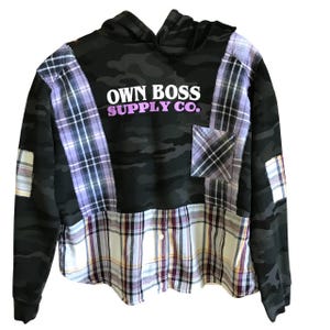 May include: A black camouflage hoodie with patchwork plaid accents. The hoodie features the text "OWN BOSS SUPPLY CO." in white and purple. The design incorporates a pocket and patches of purple, white, and black plaid fabric.