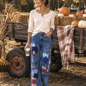May include: A long-sleeve cream-colored henley shirt with a button-front placket paired with blue denim jeans featuring various rectangular patches in shades of blue, red, and plaid patterns, finished with a raw, frayed hem at the bottom.